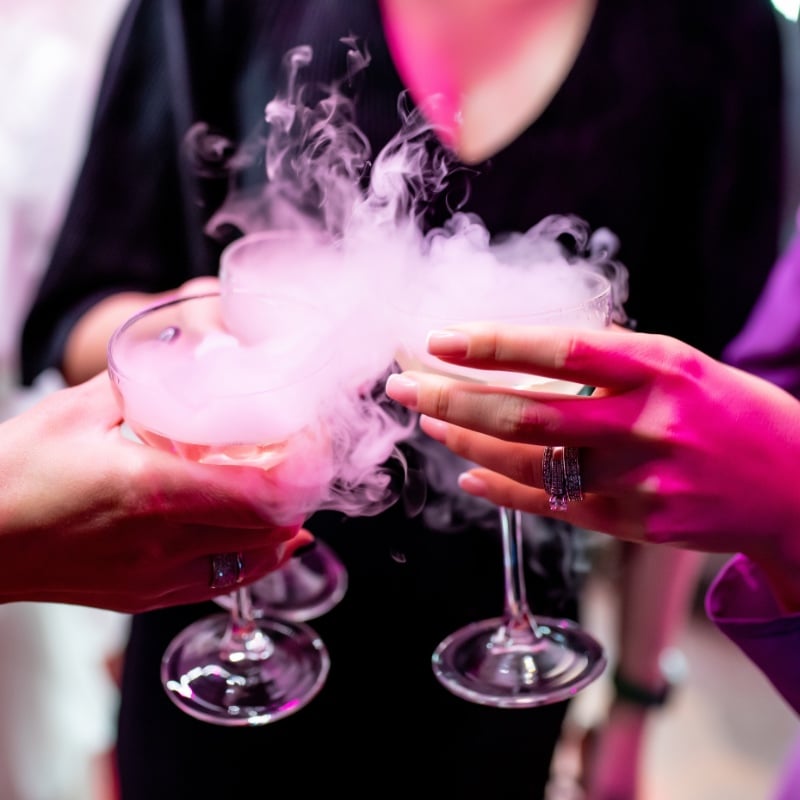 Hands of three girls holding glasses with champagne and dry ice. Beautiful white steam from glasses