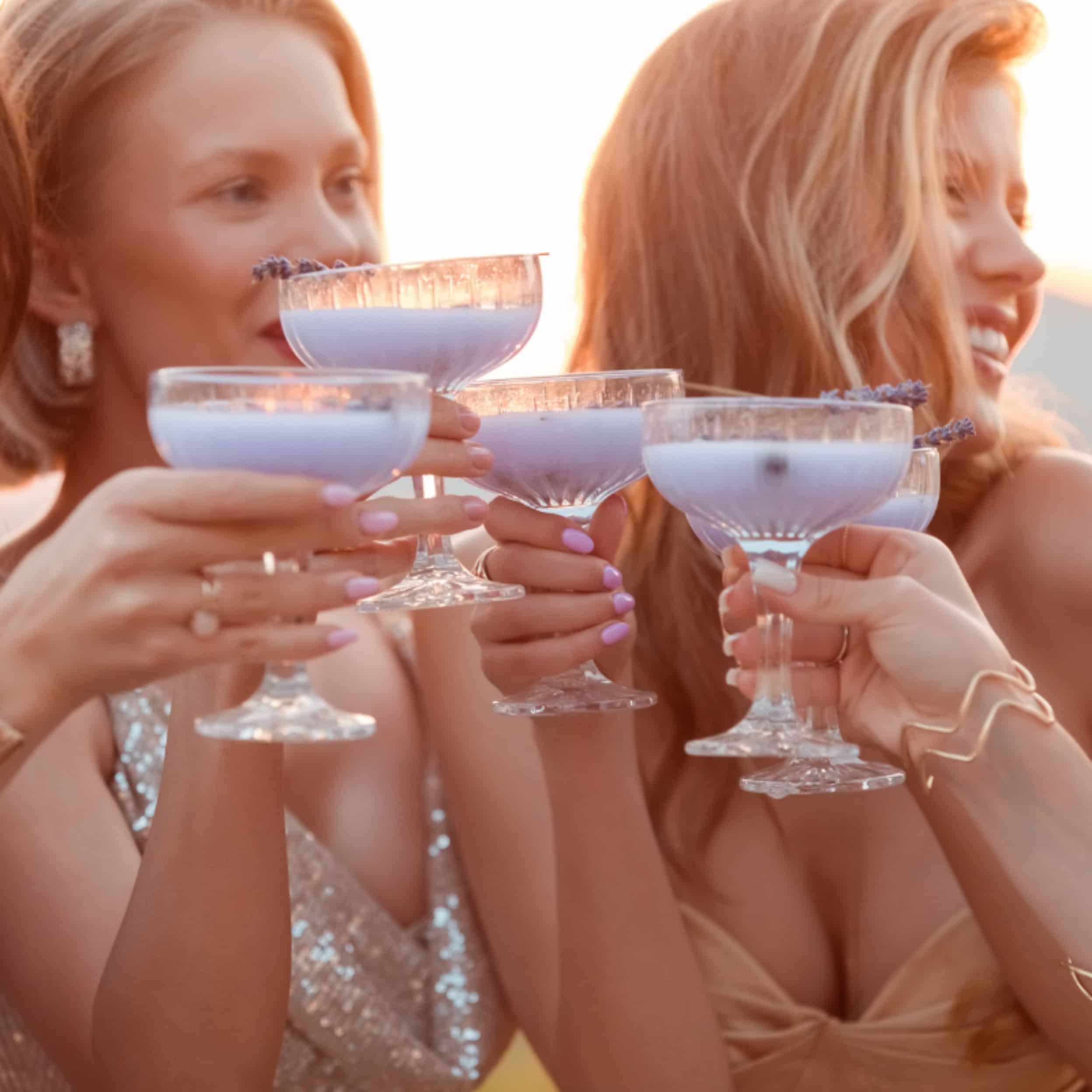 Female friends wearing elegant dresses drinking lavender cocktails at wedding reception